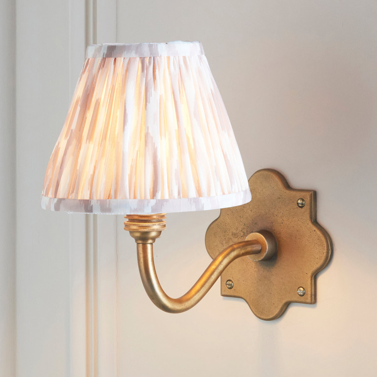 Anvil Curve brass wall light lit on a panelled wall, pale pleated ikat shade glowing warmly