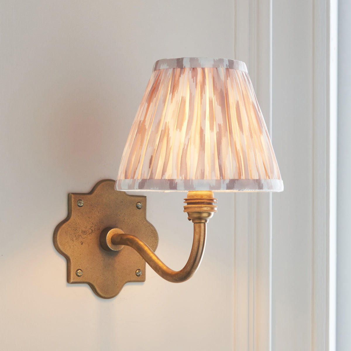 Anvil Curve antique brass wall light angled on panelling, natural ikat pleated shade illuminated