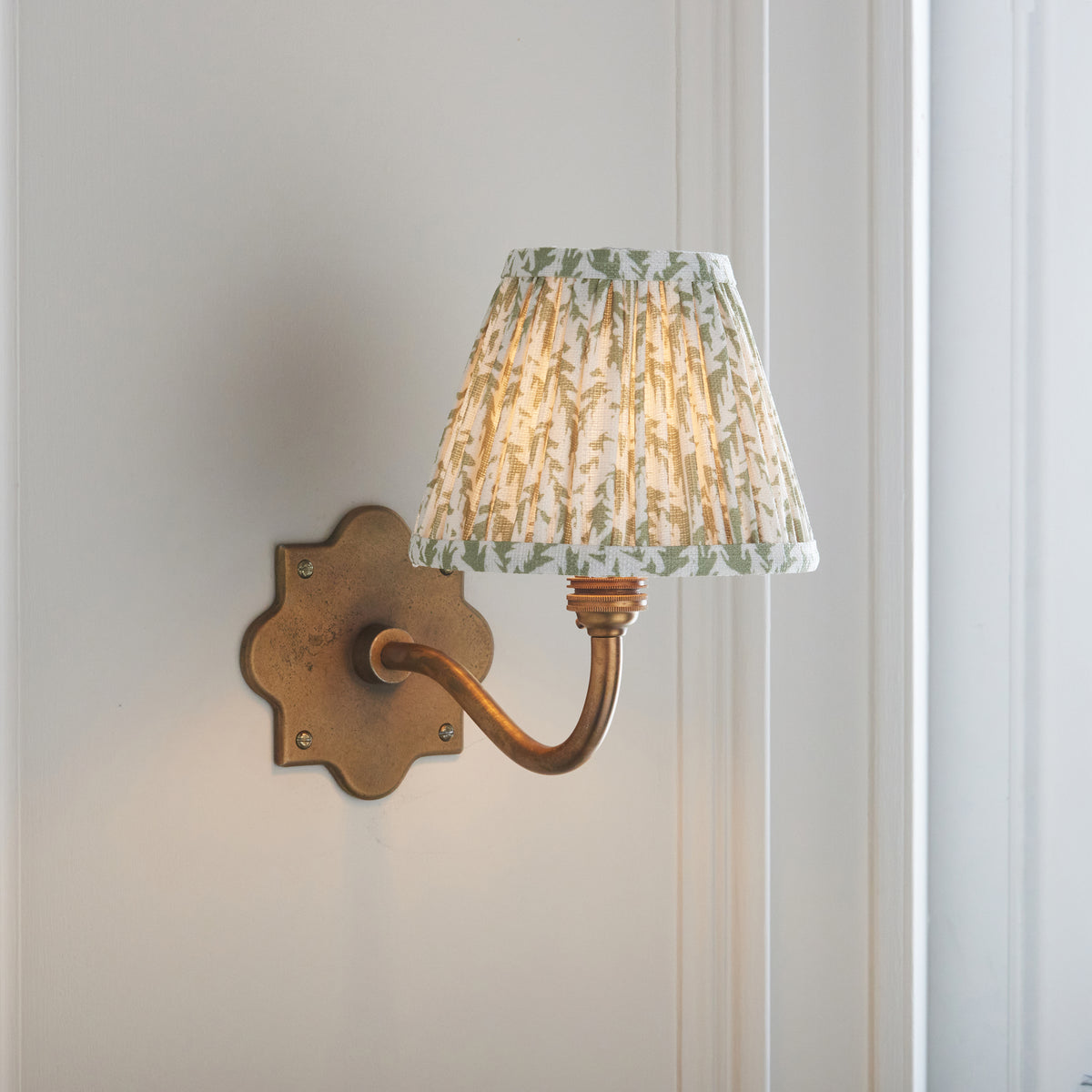 Anvil Curve antique brass wall light on panelling, angled view of its green foliage pleated shade
