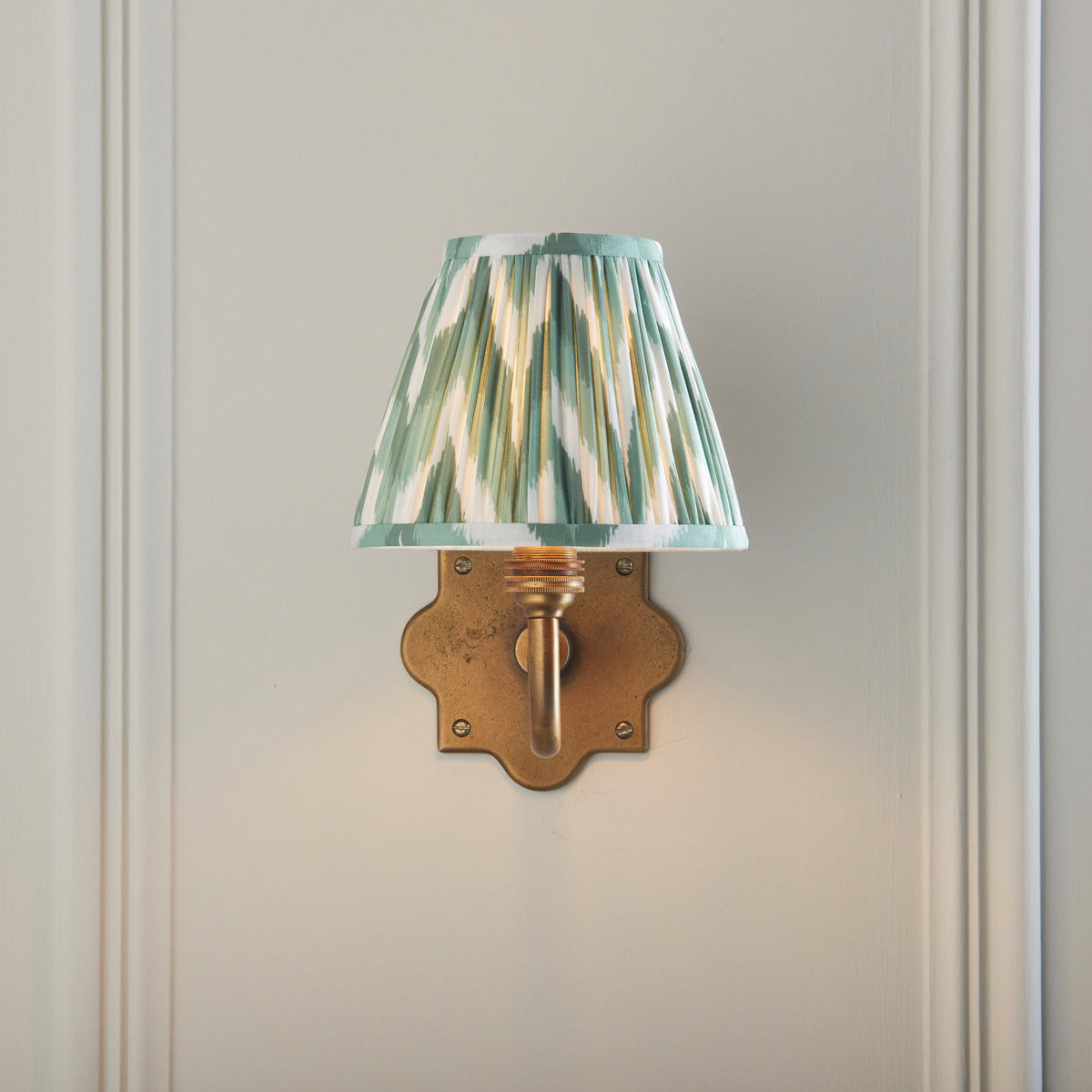 Front view of the Anvil Curve wall light, lit green zigzag ikat shade above a quatrefoil brass plate