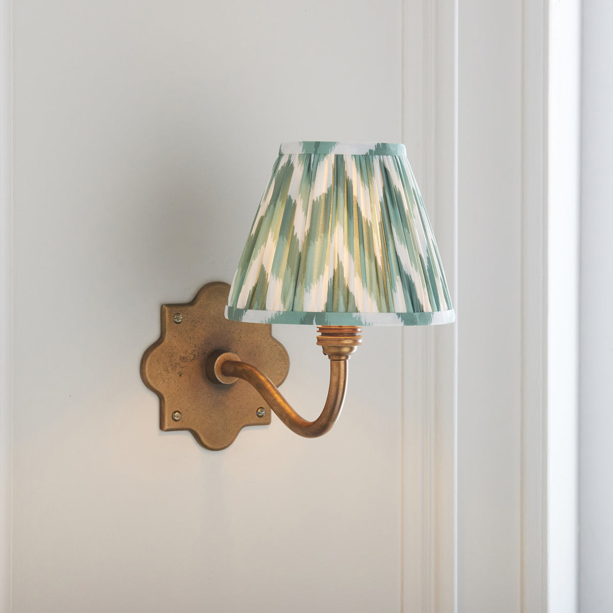 Anvil Curve antique brass wall light angled on panelling with sage zigzag ikat pleated shade