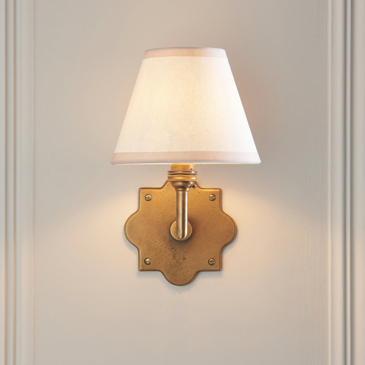 Front view of the Anvil Angle wall light, ivory tapered shade above a quatrefoil brass backplate