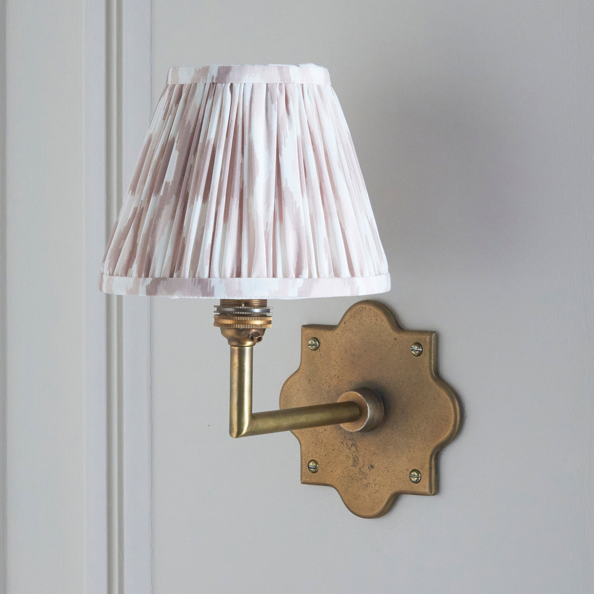 Anvil Angle wall light on pale panelling, natural ikat pleated shade lit above a brass arm
