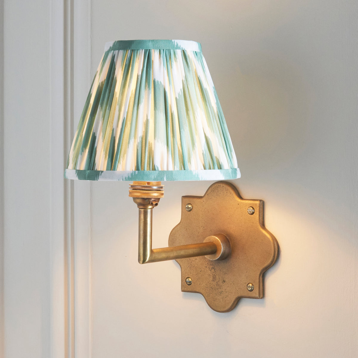 Anvil Angle brass wall light lit on a panelled off-white wall, green ikat pleated shade angled left