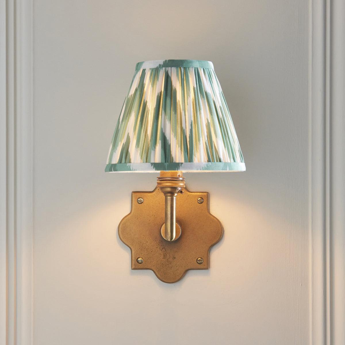 Anvil Angle brass wall light shown head-on, glowing green ikat shade above a star-shaped brass backplate