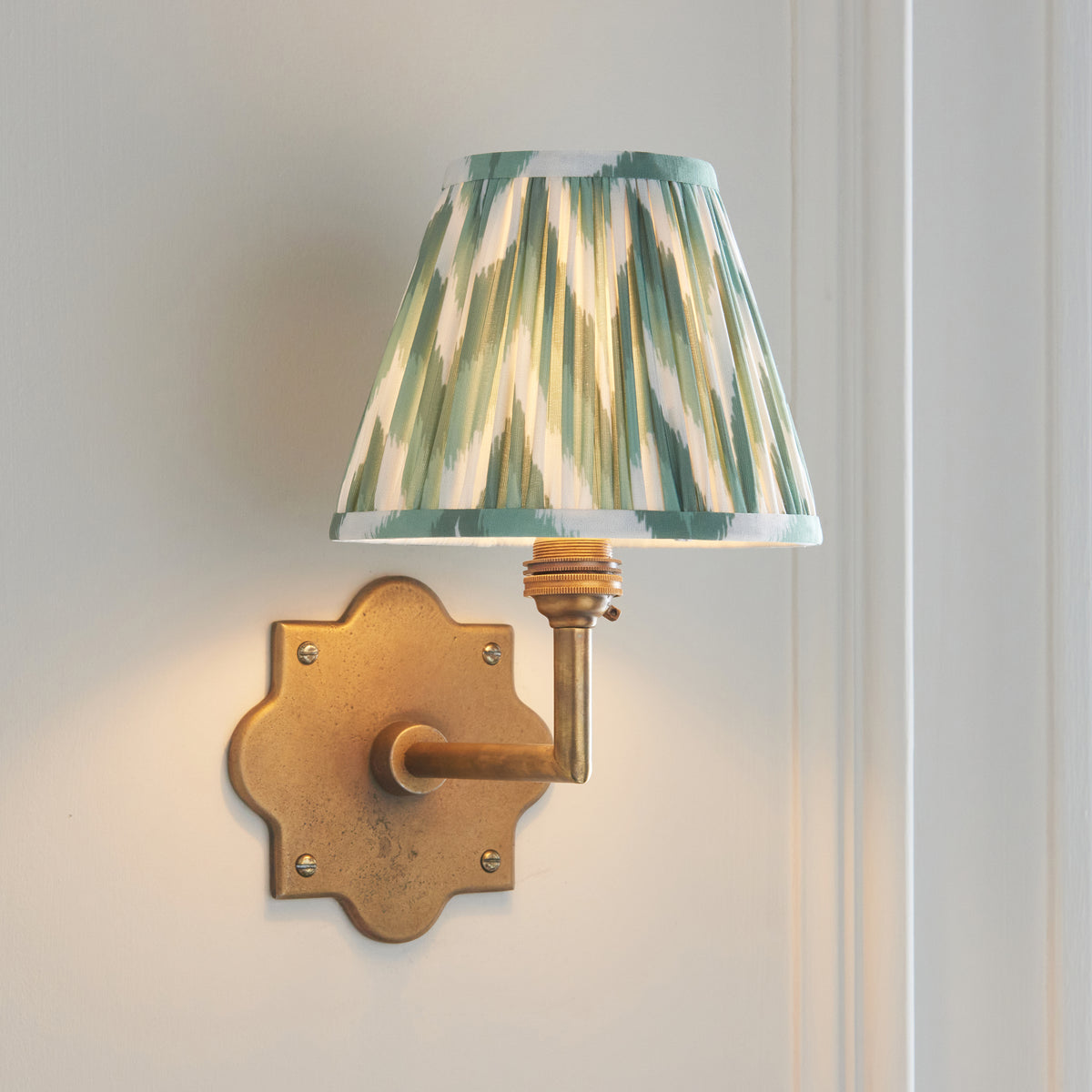 Anvil Angle brass wall light lit against pale panelling, pleated green ikat shade casting a warm glow
