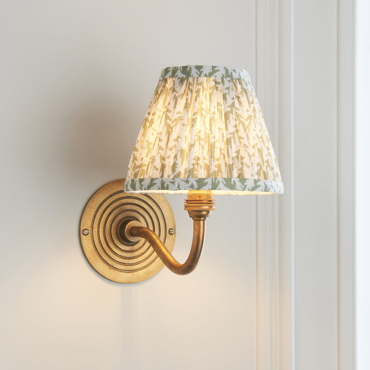 Ripple Curve & Leaf antique brass wall light angled on pale panelling with warm leaf-print shade