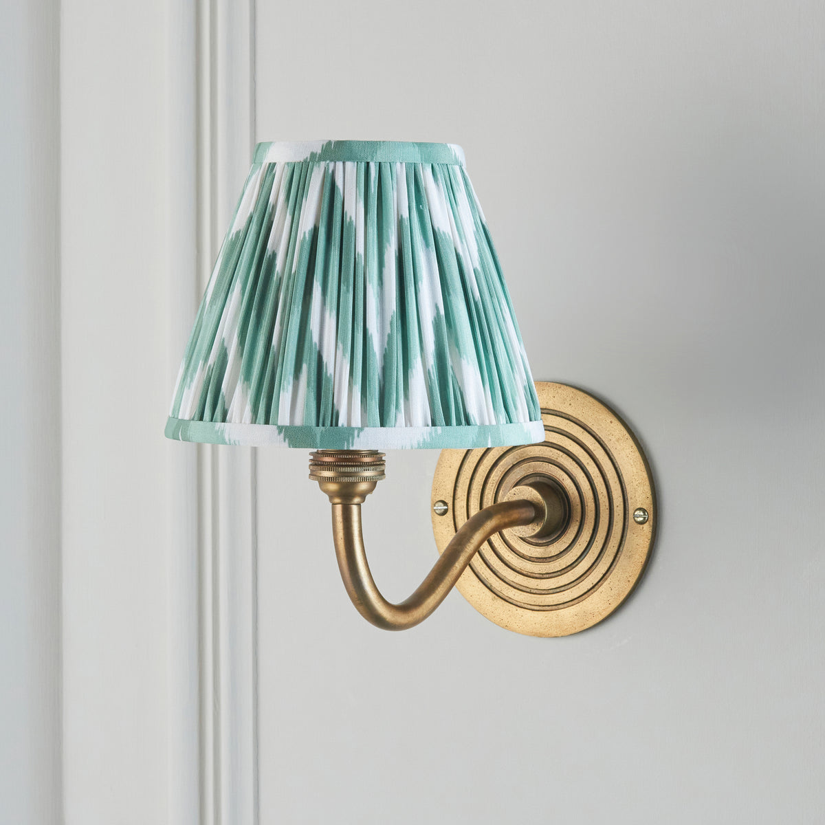 Ripple Curve brass wall light on a grey panelled wall, softer green ikat pleated shade illuminated