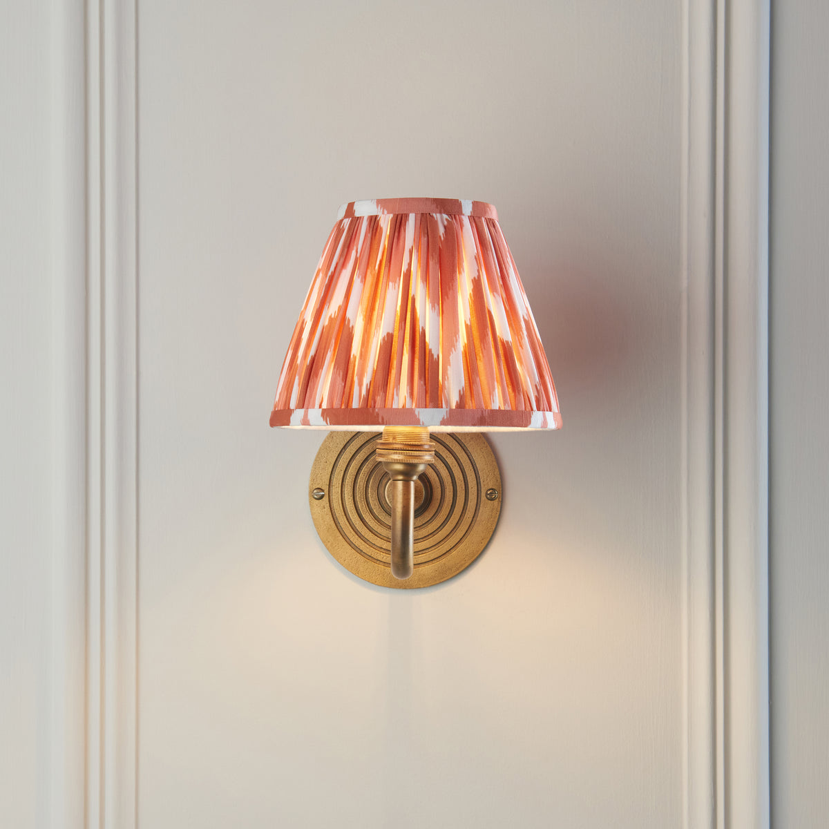 Ripple Curve brass wall light head-on against pale panelling with warm orange ikat shade