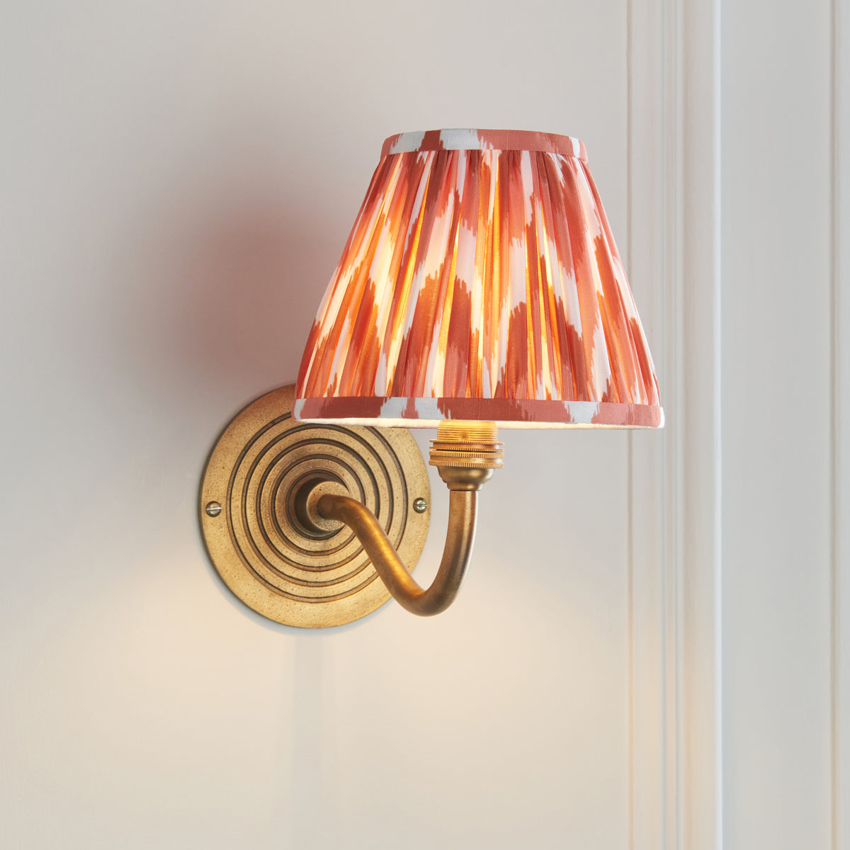 Ripple Curve antique brass wall light angled on off-white wall, pleated orange ikat shade lit