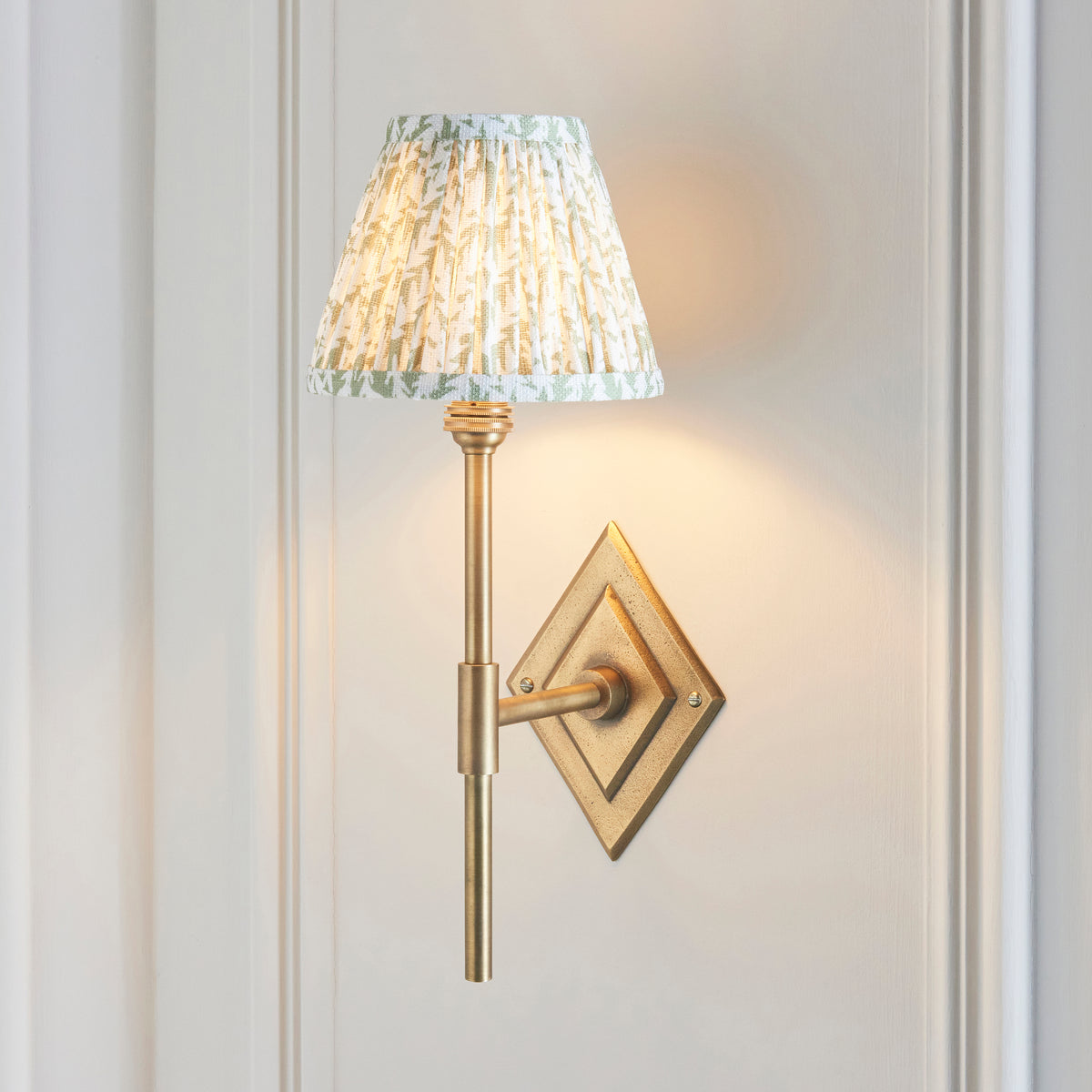 Diamond Tall brass wall light lit on panelled off-white wall, pleated leaf-print shade glowing