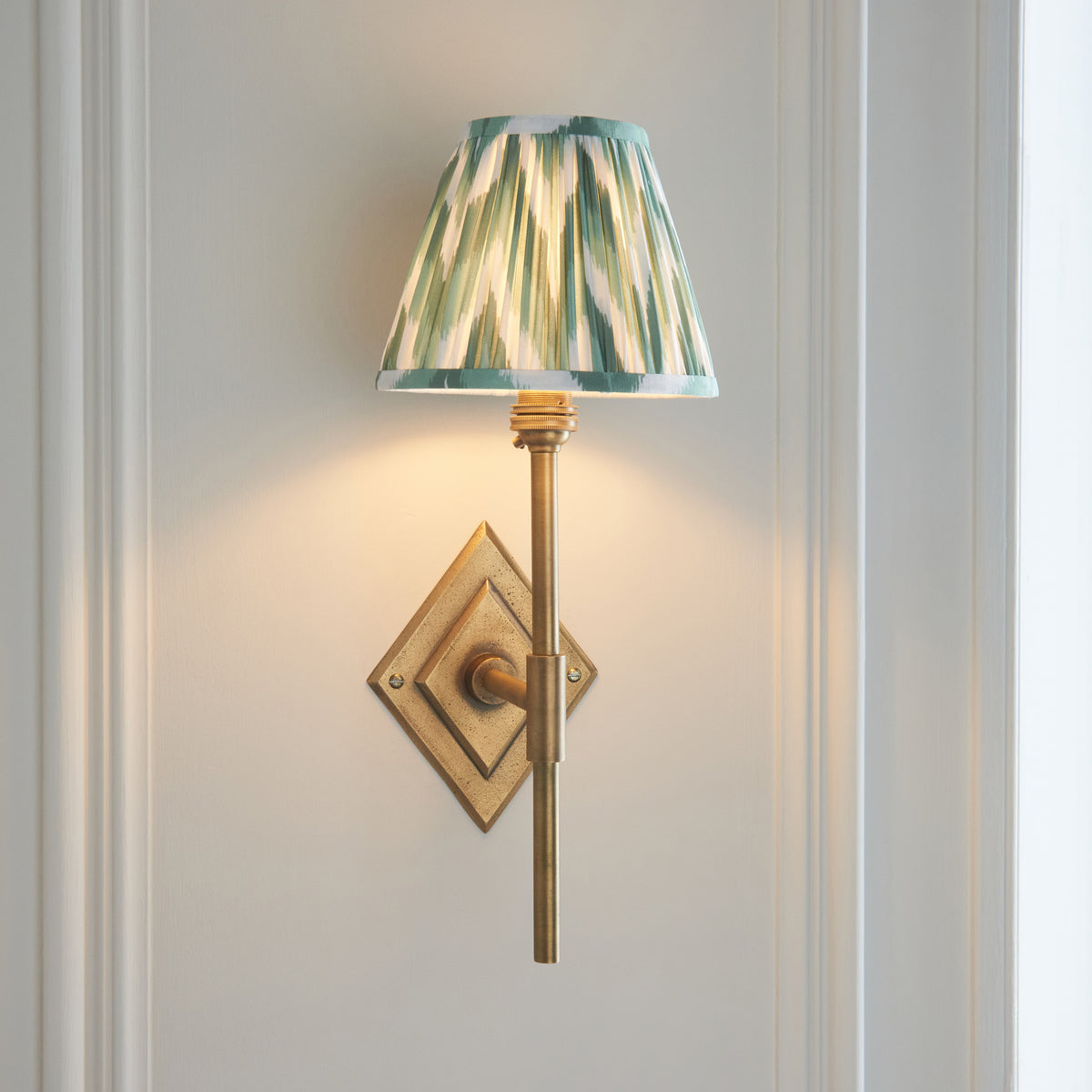 Diamond Tall antique brass wall light angled on off-white panelling with lit green ikat shade