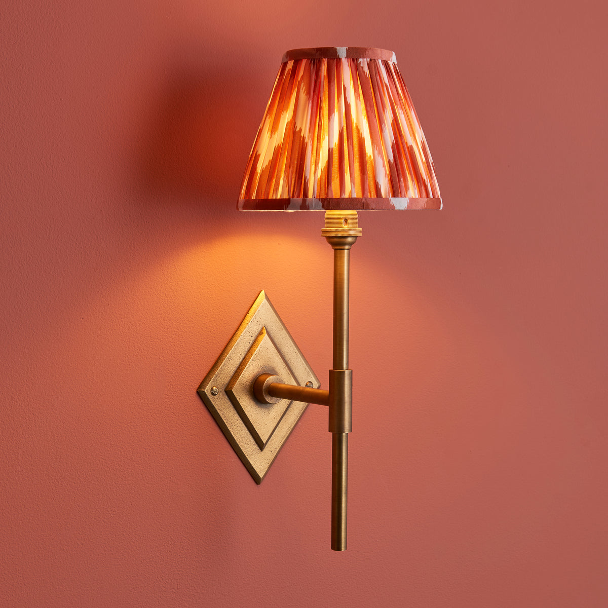 Diamond Tall brass wall light on a terracotta wall, glowing orange ikat pleated shade