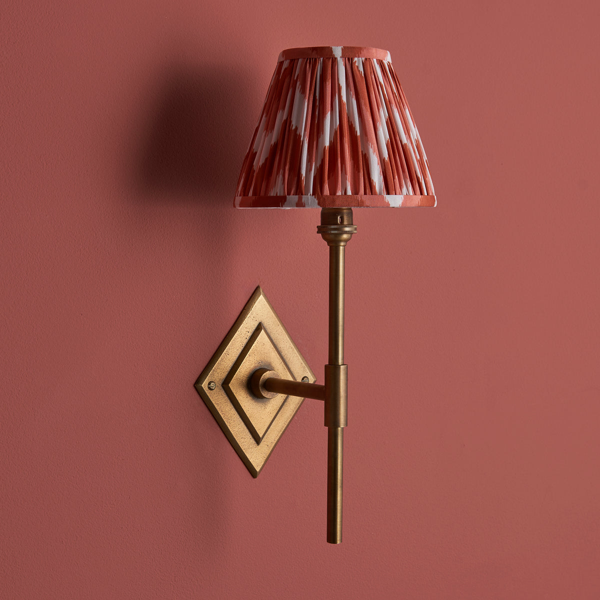 Diamond Tall brass wall light on a deep terracotta wall, russet ikat pleated shade casting shadow