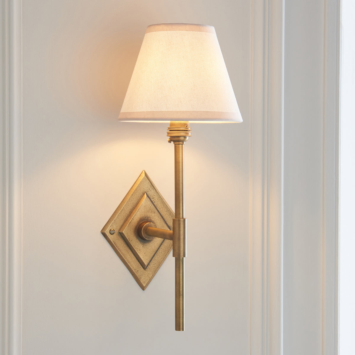 Diamond Tall antique brass wall light angled on off-white panelling with lit white fabric shade