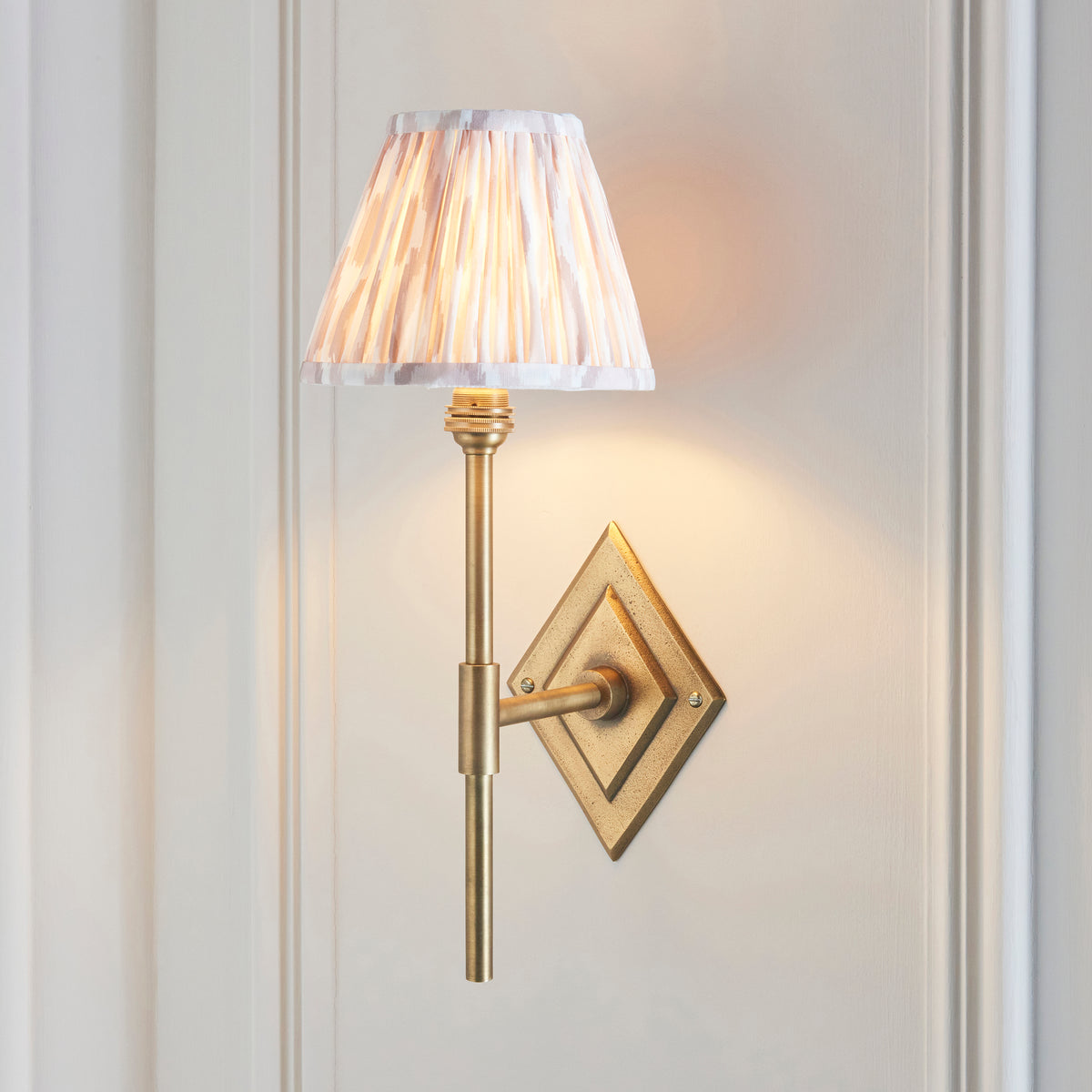 Diamond antique brass wall light lit on a panelled wall, natural ikat pleated shade glowing softly