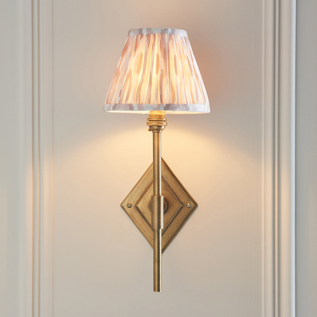 Front view of the Diamond antique brass wall light on a cream wall, ikat pleated shade illuminated