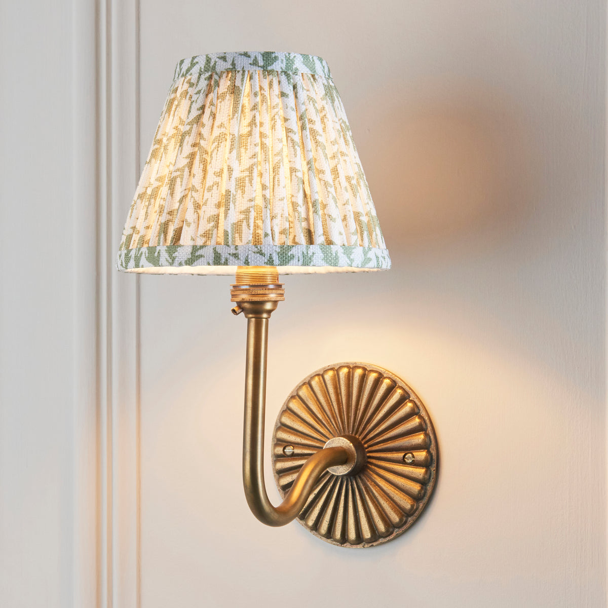 Dahlia Swan brass wall light lit on a panelled wall, green leaf-print pleated shade glowing