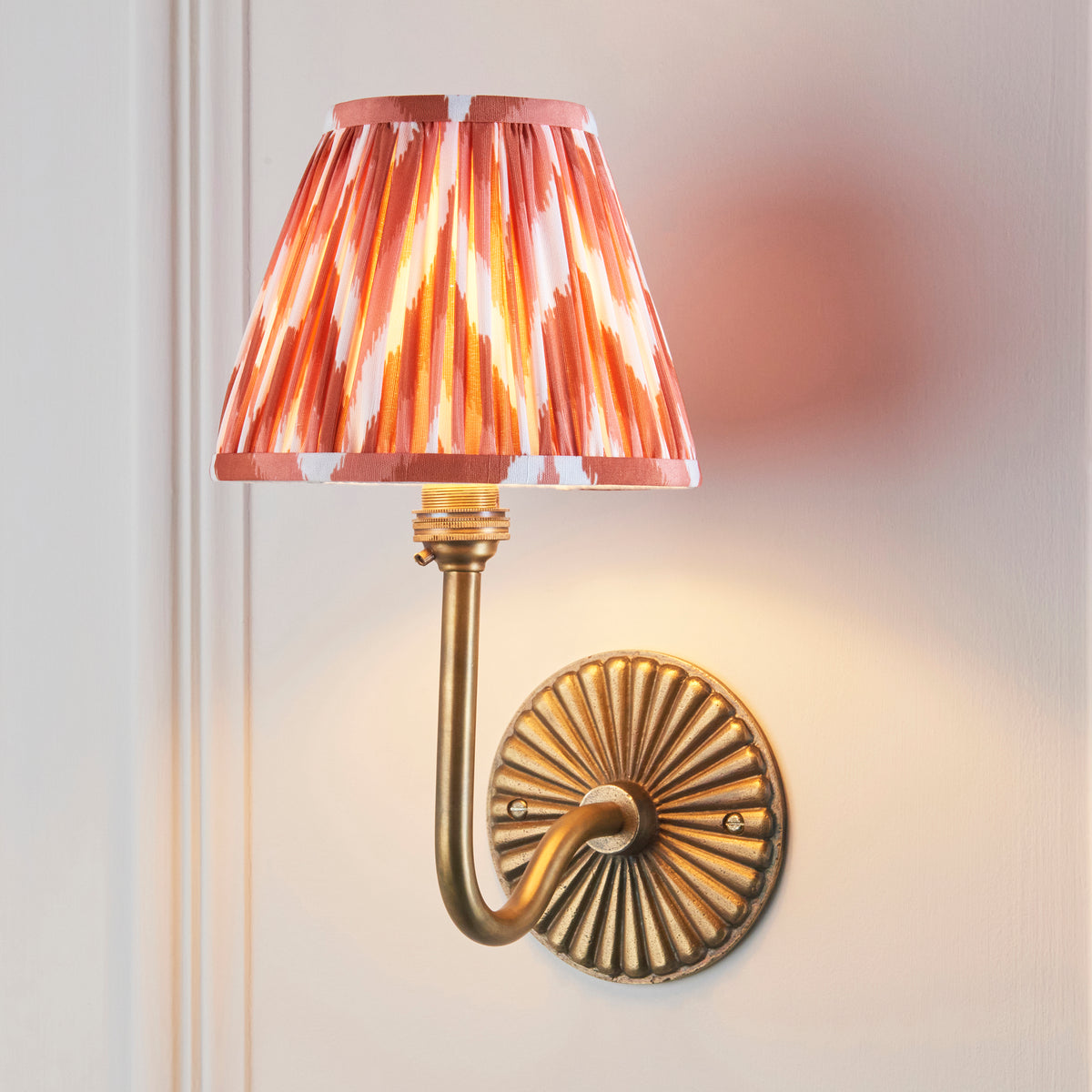 Dahlia Swan brass wall light lit on a panelled wall, warm orange ikat pleated shade glowing