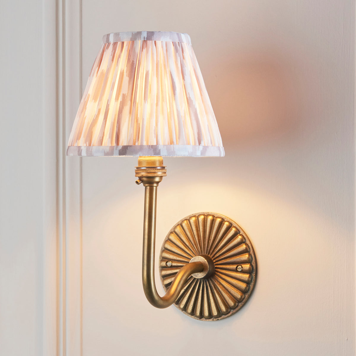 Dahlia Swan brass wall light lit on panelled wall, natural beige ikat pleated shade glowing