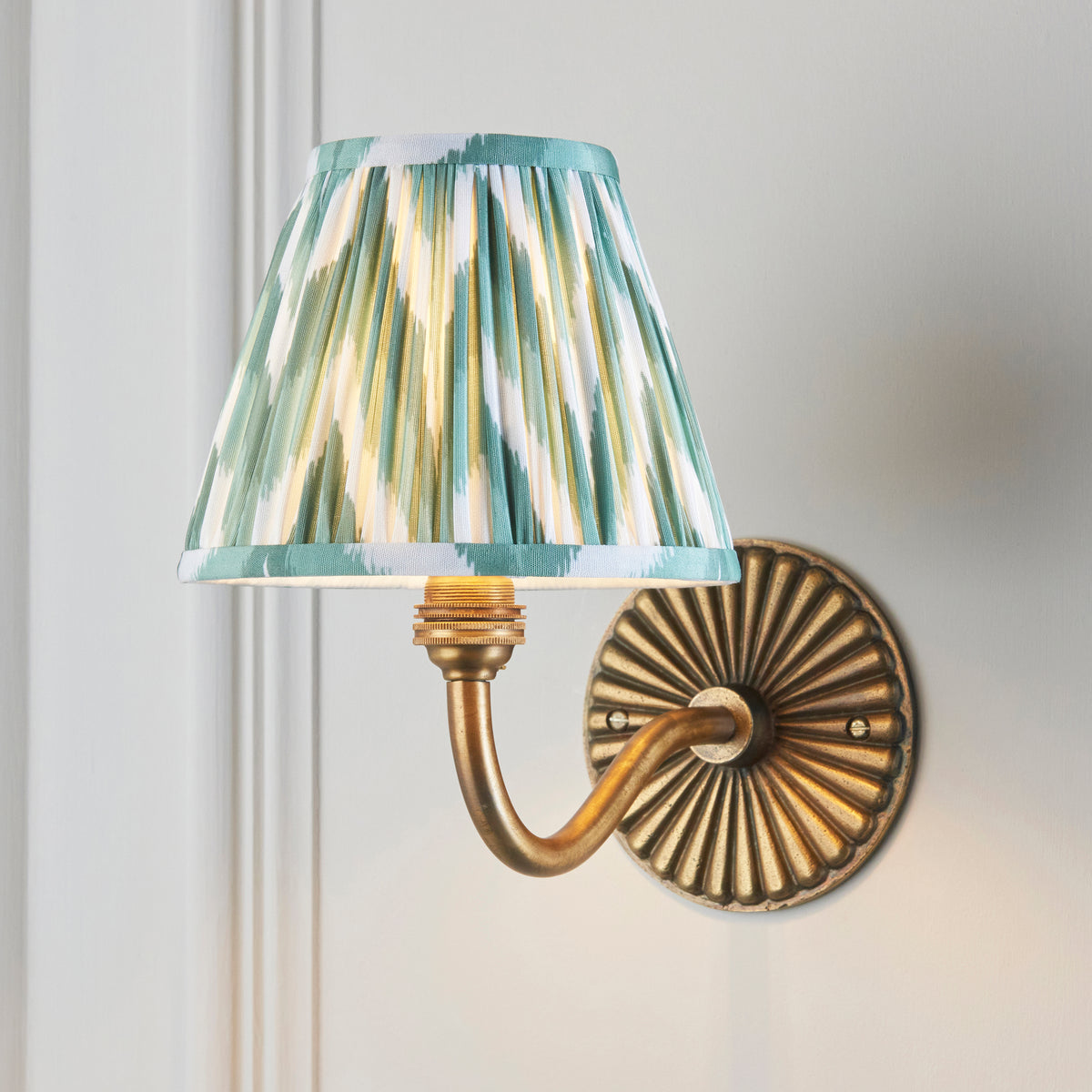 Dahlia Curve brass wall light lit on panelled wall, green zigzag ikat pleated shade glowing