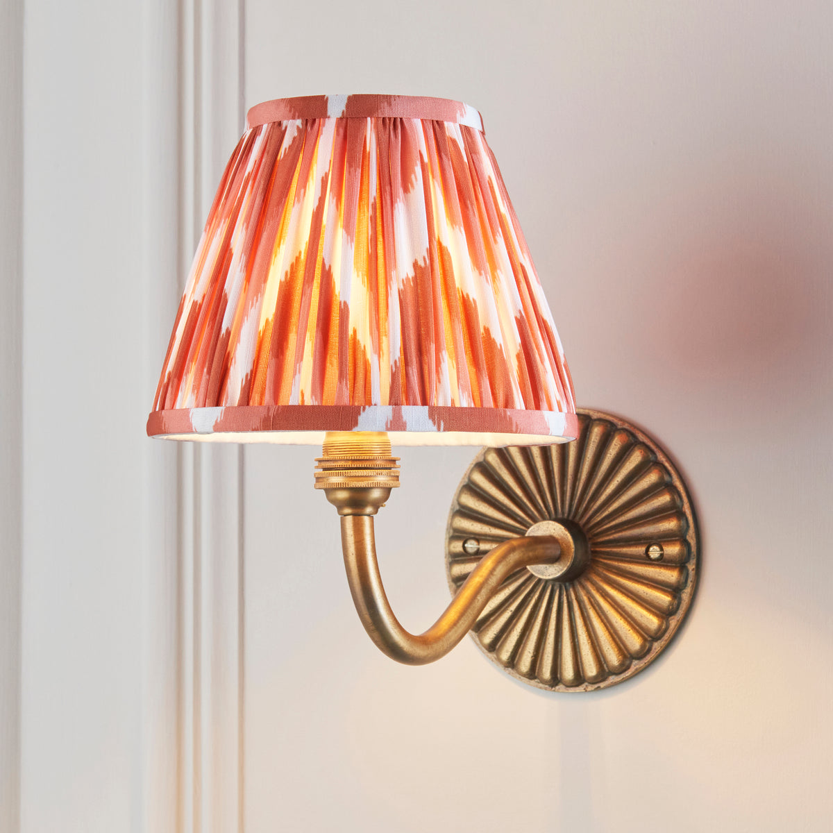 Orange ikat pleated shade lit on the Dahlia Curve brass wall light, mounted on a pale panelled wall