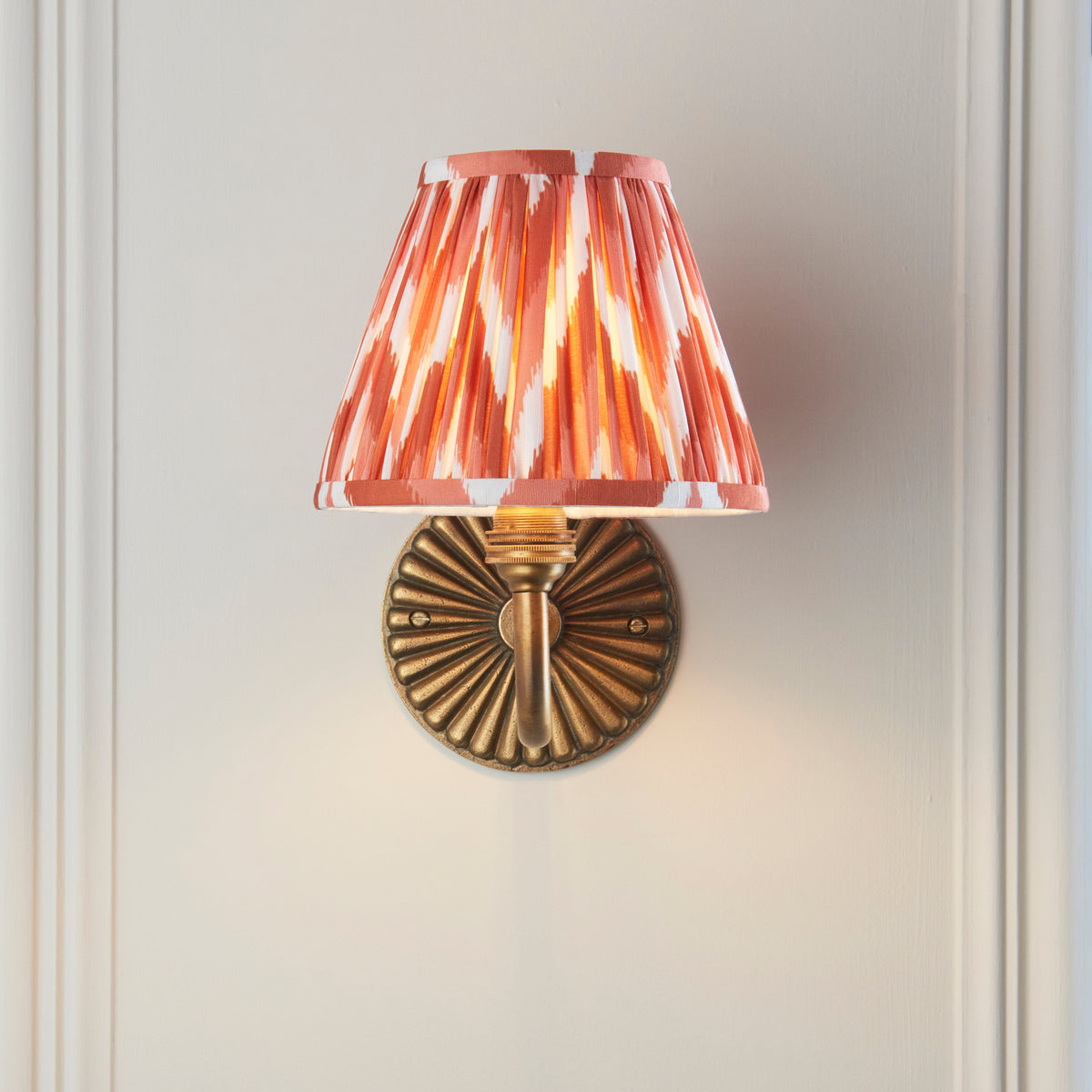 Front view of the Dahlia Curve wall light, its ribbed brass backplate below a glowing orange zigzag shade