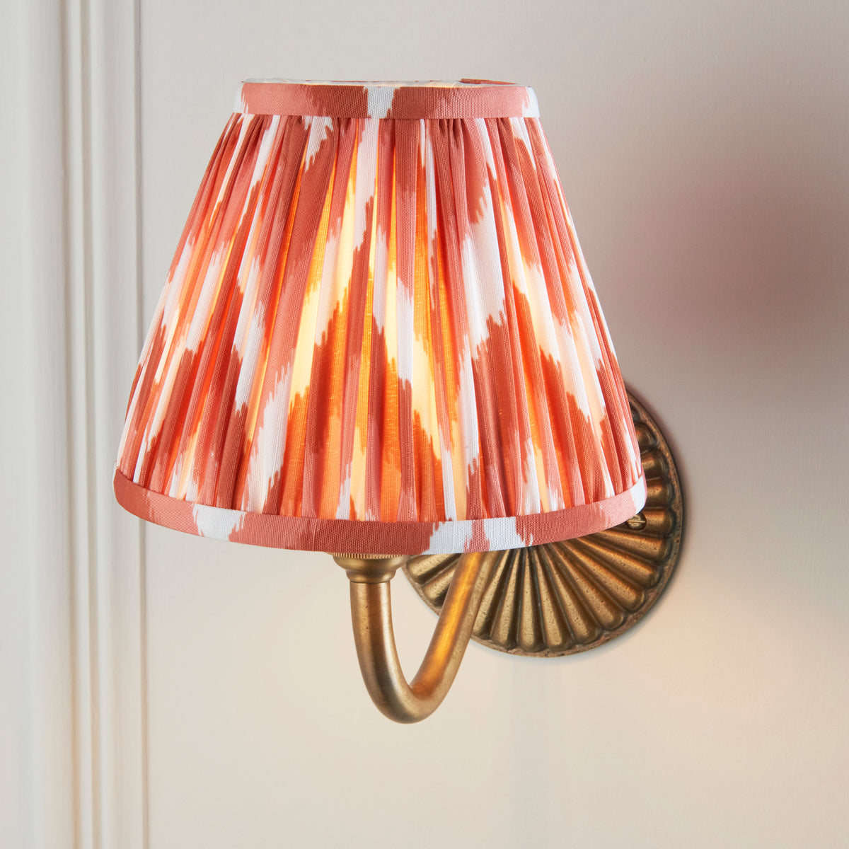 Close view of the illuminated orange and white pleated ikat shade above the curved brass sconce arm