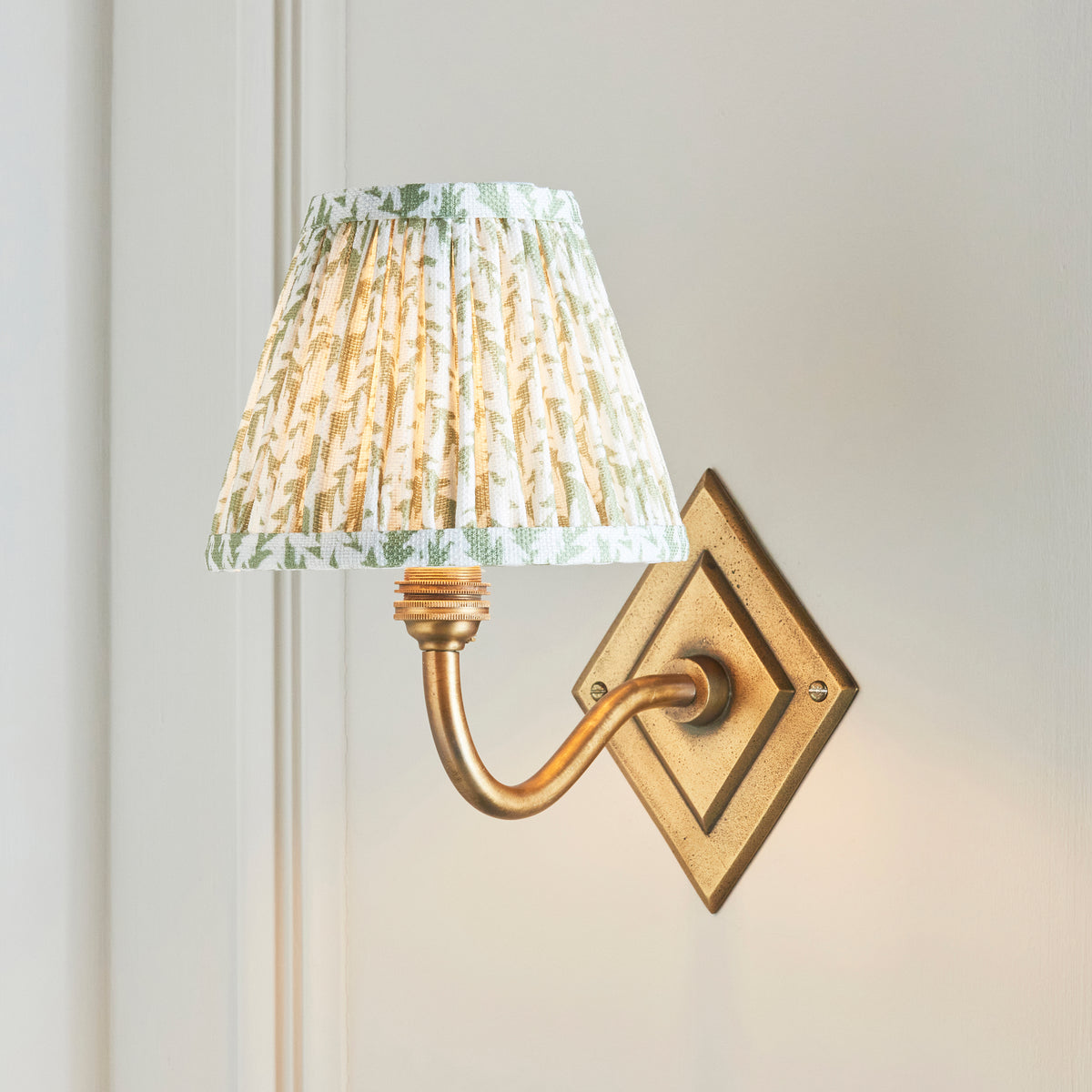 Green leaf pleated shade lit on the Diamond Curve brass wall light against pale panelling