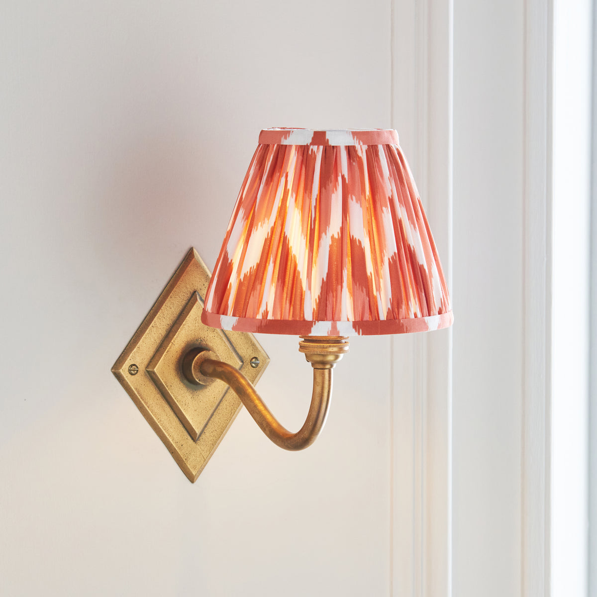 Angled view of the Diamond Curve brass wall light with warm orange and white pleated shade