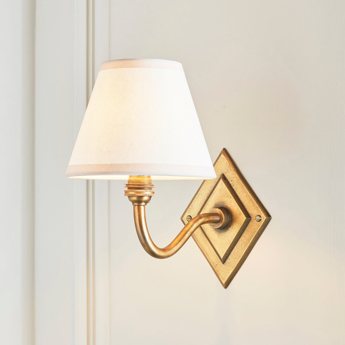 White fabric shade lit on the Diamond Curve brass wall light against pale wall panelling