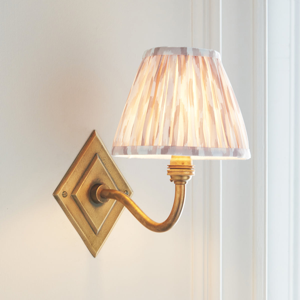 Angled view of the Diamond Curve brass wall light with warm-lit beige ikat pleated shade