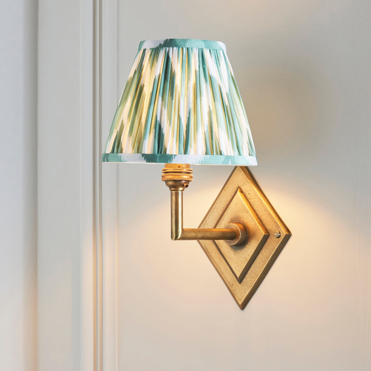 Diamond Angle brass wall light with green ikat pleated shade, angled against white panelling