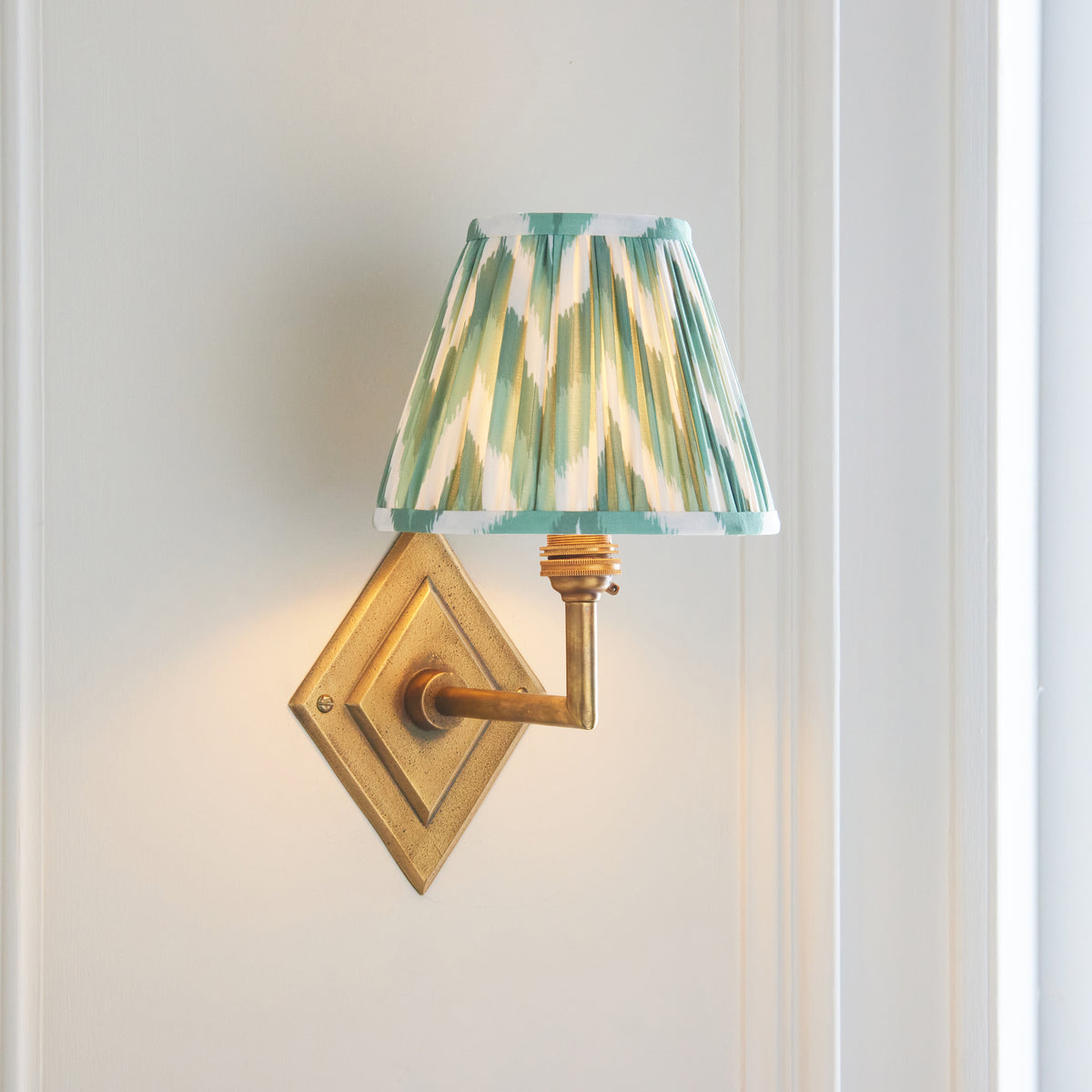Diamond Angle brass wall light glowing warmly on a white panelled wall, green ikat shade