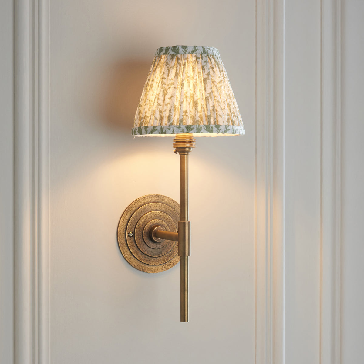 Halo brass wall light casting warm light on a white panelled wall, leaf-print shade