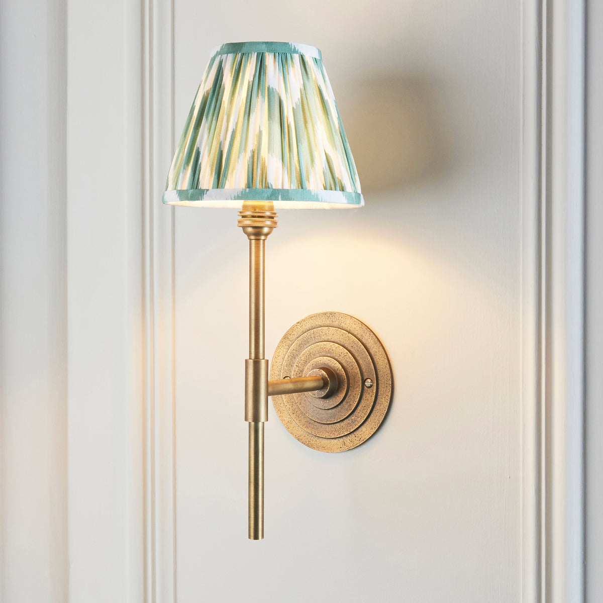 Halo Tall brass wall light with green ikat shade, lit and angled on white panelling