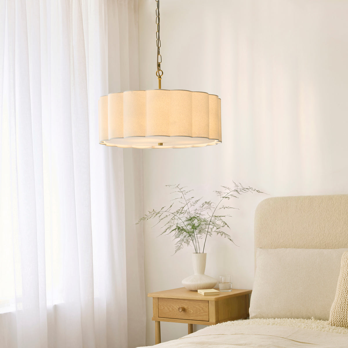 Serene brass pendant lit above a bedside table with vase of foliage and cream upholstered bed