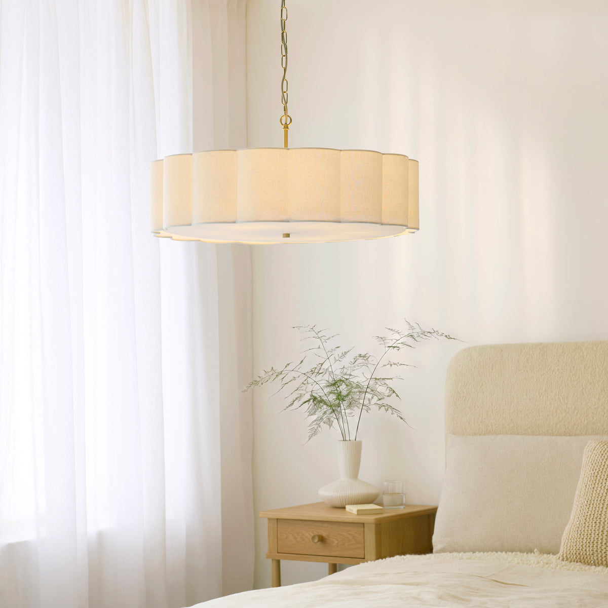 Serene brass chandelier lit above a bedside table with foliage vase and cream bed