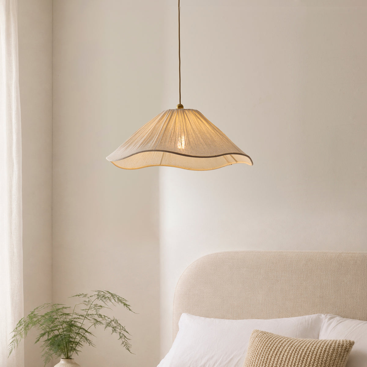 Isabella pleated white pendant lit above a bed with cream headboard and fern in a bedroom