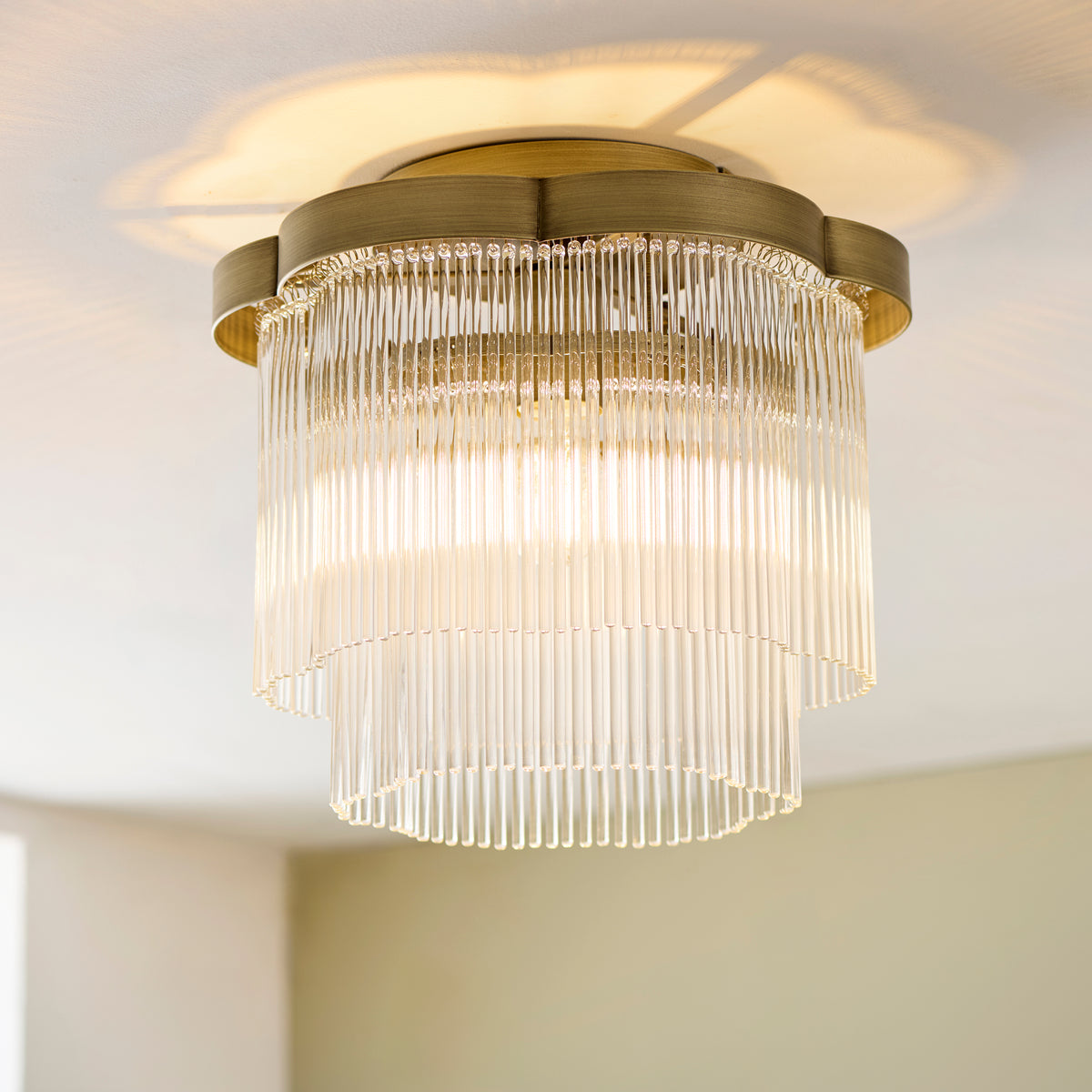 Angled view of the lit Lucille antique brass flush light showing its tiered clear glass rod shade