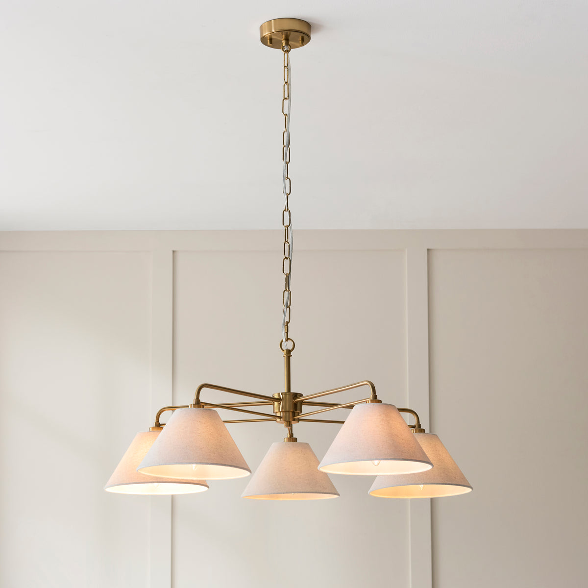 Lit Wallbridge pendant with five tapered linen shades hanging from an aged brass frame