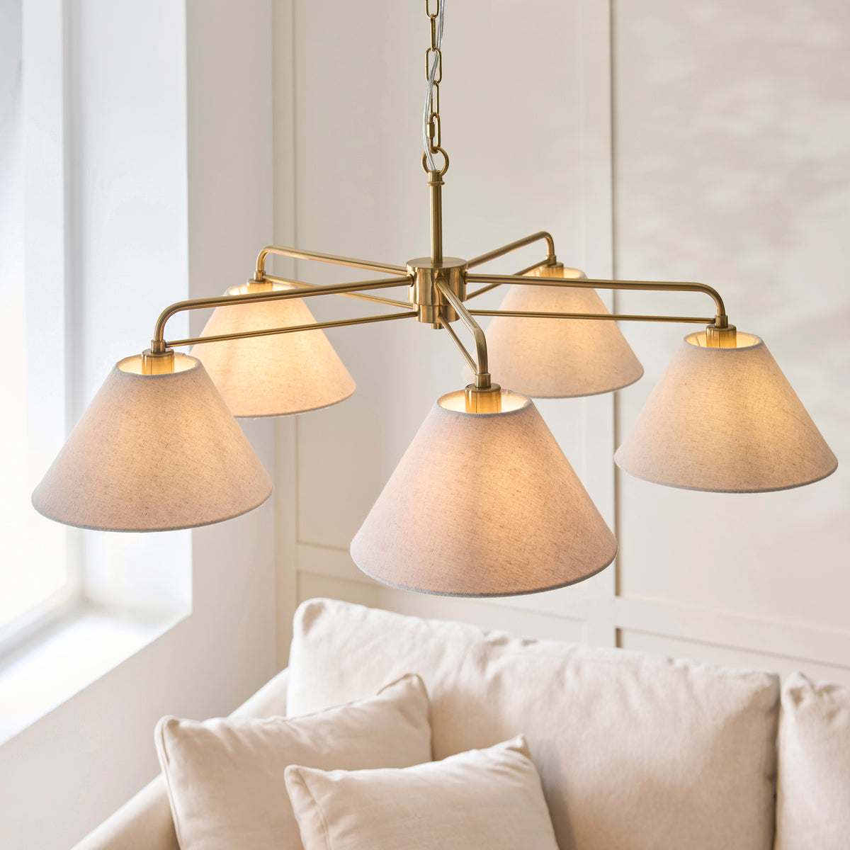 Wallbridge brass pendant lit beside a bright window above a cream sofa with cushions