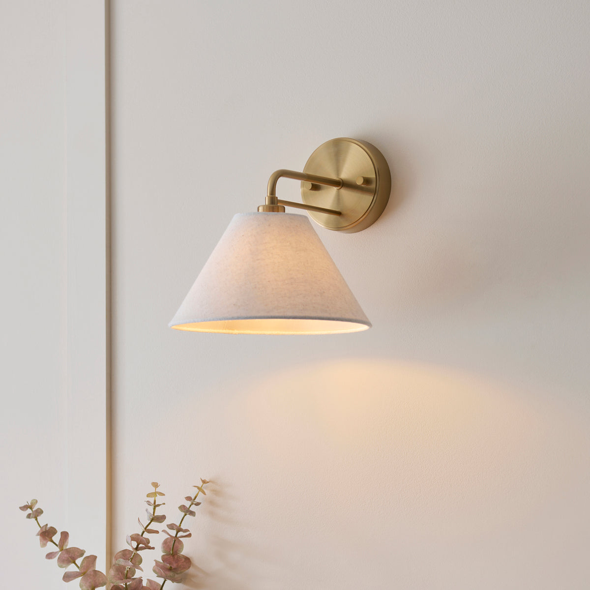 Wallbridge brass wall light casting warm light on a pale wall beside dried eucalyptus stems