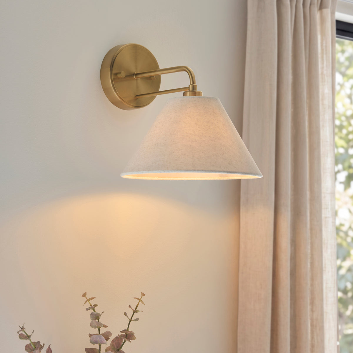 Wallbridge wall light lit on a neutral wall next to linen curtains and eucalyptus foliage