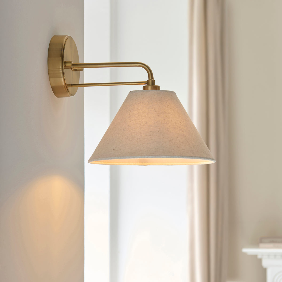 Wallbridge brass wall light angled from a pale wall, glowing linen shade above a mantelpiece