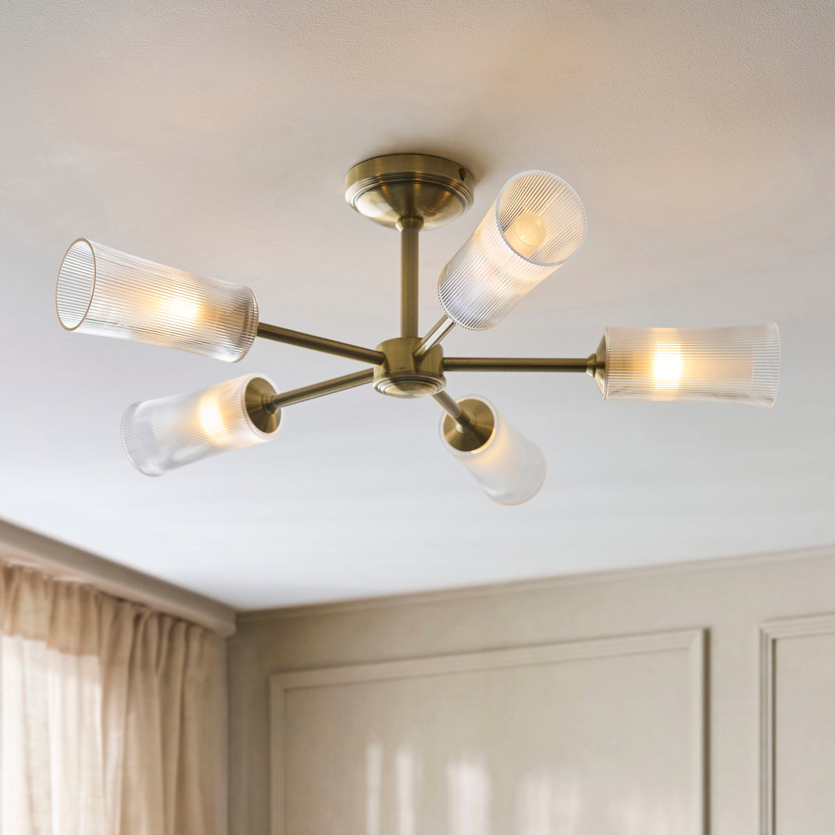 Avelyn five-arm brass semi flush light lit beside pale curtains and fitted wardrobes