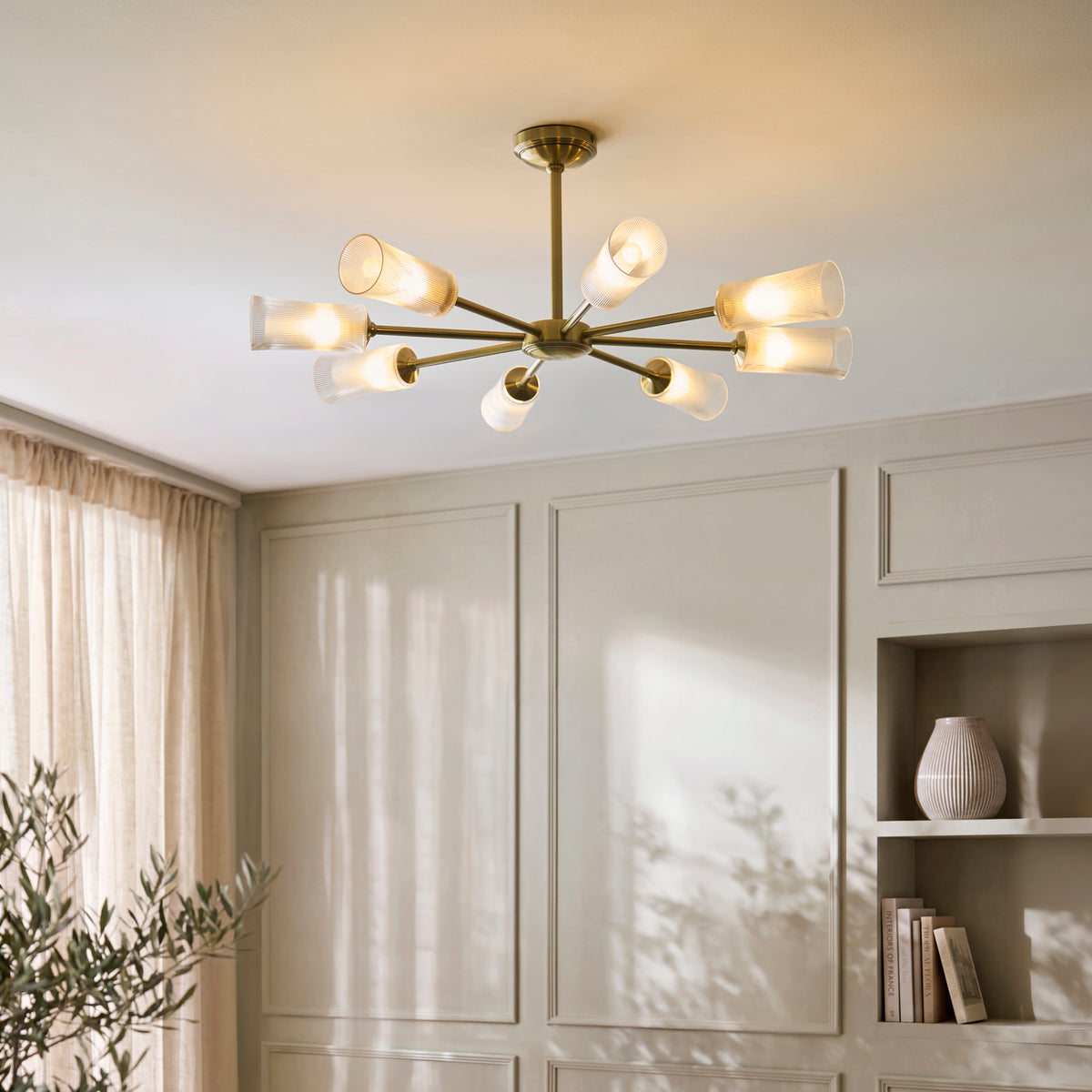 Avelyn brass eight-light chandelier lit below a ceiling in a panelled room with shelving