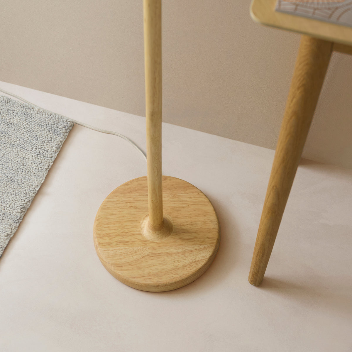 Round oak-stained wooden base of the Olson floor lamp on a pale floor beside a grey rug and table leg
