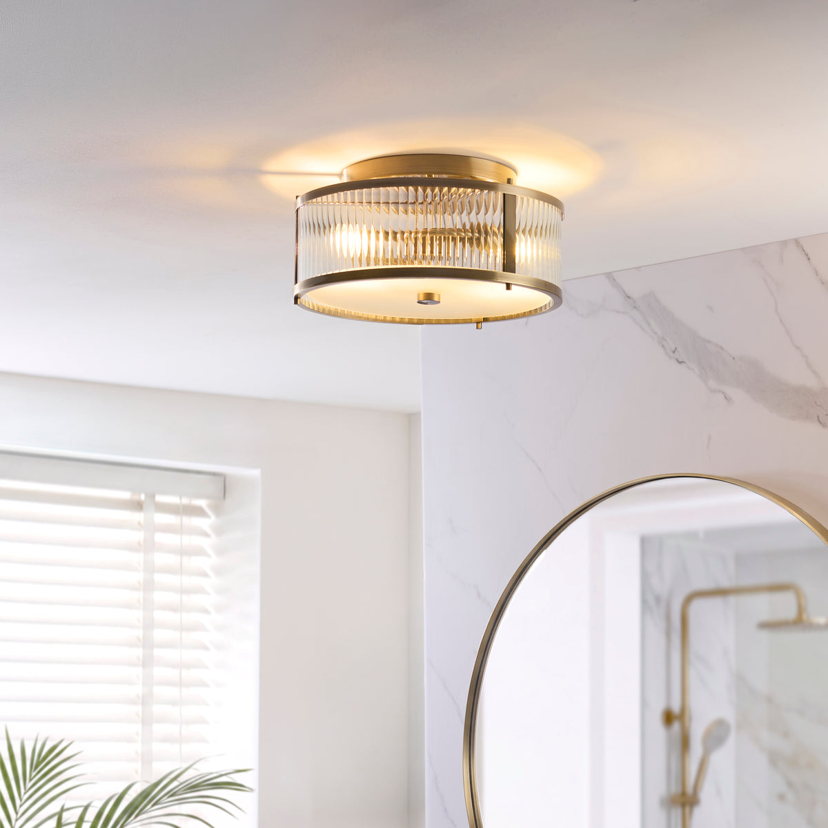 Ridgeton brass flush ceiling light lit in a marble bathroom with round mirror and shower