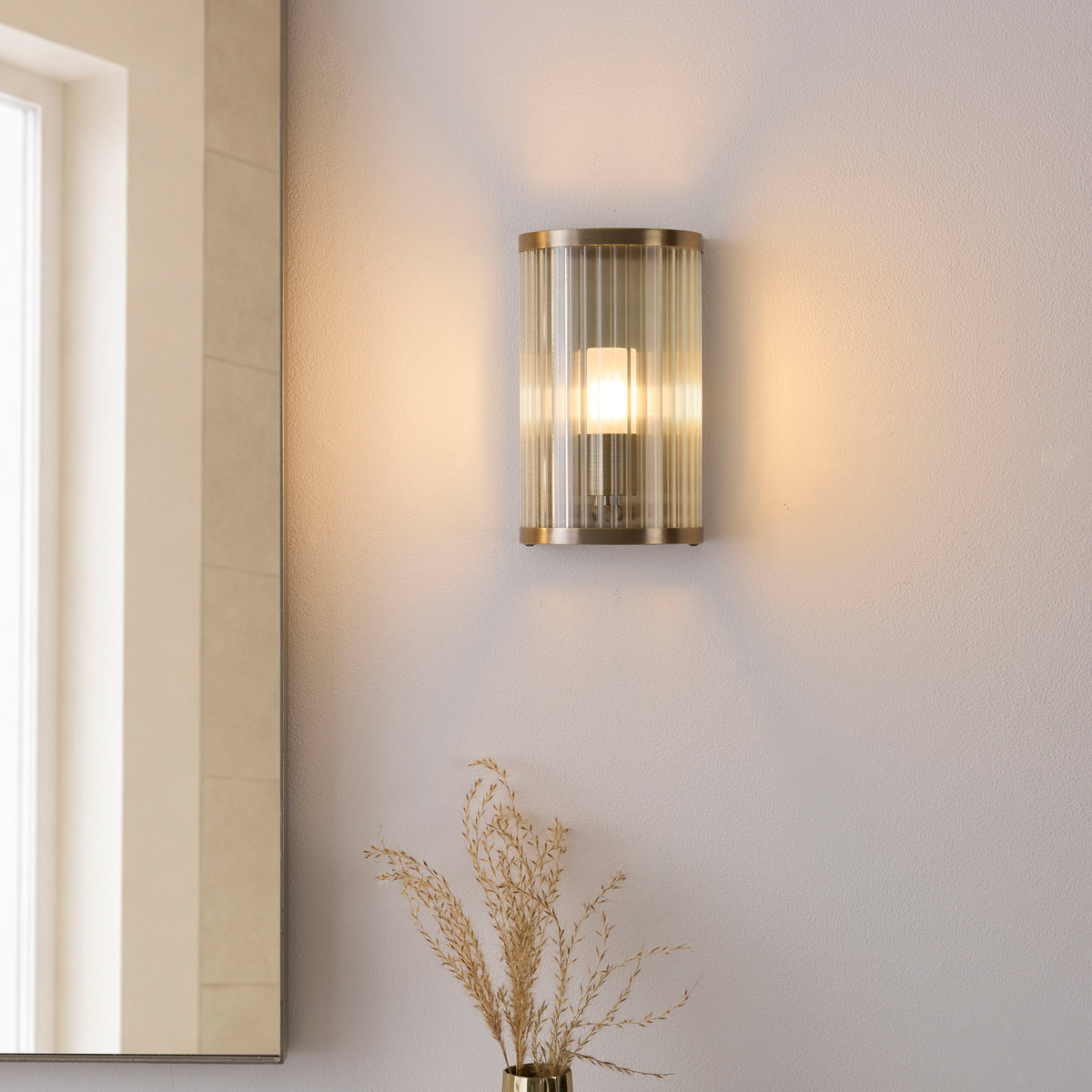 Ridgeton ribbed glass wall light glowing on a bathroom wall beside a mirror and dried grasses